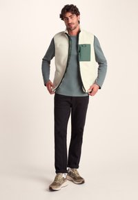 Fleece vest in cream with green accents, featuring a zippered front and chest pocket. Worn over a long-sleeve teal top and black pants.