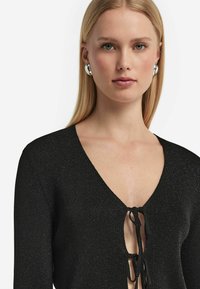 Svart stickad tröja med V-ringning, som har en dekorativ knytning vid bröstet och en subtil glänsande struktur genom hela tyget.