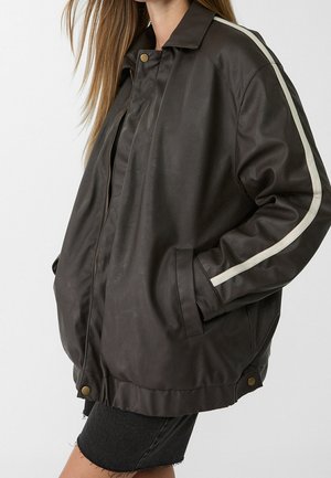 Woman wearing a dark brown leather jacket with a white stripe on the sleeve, side pockets, and brass snap buttons.