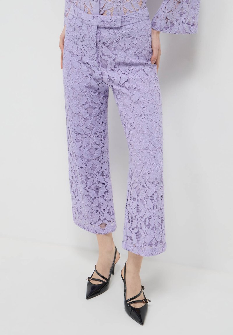 Un pantalon large en dentelle violette présente des motifs floraux délicats, une taille haute et une longueur raccourcie, associé à des talons en cuir verni noir.