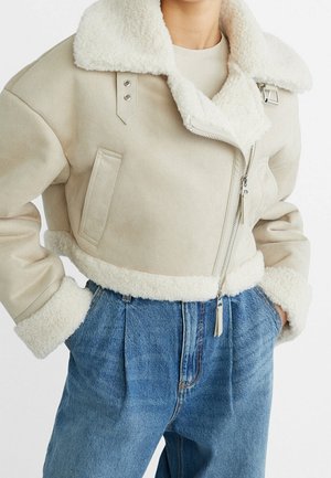 Beige verkürzte Lammfelljacke mit großem Kragen und Reißverschluss, kombiniert mit hoch taillierten blauen Jeans, Hände in den Taschen.