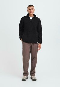 Sudadera negra de forro polar con diseño de media cremallera, que presenta una superficie texturizada. Combinada con pantalones marrones y calzado gris.
