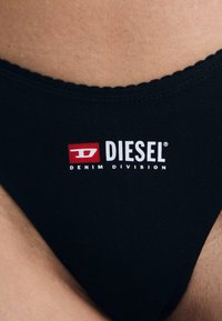 Zwarte bikini onderkant met een golvende tailleband, voorzien van een rood-witte "DIESEL DENIM DIVISION" logo op de voorkant. Gladde textuur.