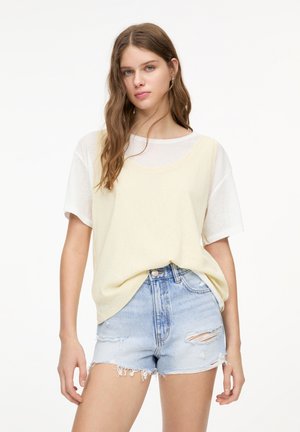 Lichtgele shirt met korte mouwen, laag over een witte top, gecombineerd met verstoorde lichtblauwe denimshorts met franjes en scheuren.
