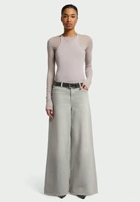 Kvinna står klädd i en åtsittande beige långärmad topp och högmidjade vida ljusgrå jeans med ett svart bälte.