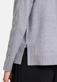 Grauer Strickpullover mit einem Kabelstrickmuster an den Ärmeln, geripptem Saum und seitlichen Schlitzen für zusätzlichen Detail- und Textur-Effekt.