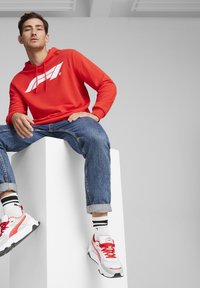 Rød hoodie med et hvidt logo, blå denimjeans og hvide sneakers med røde detaljer. Modellen sidder på et hvidt podium foran en neutral baggrund.