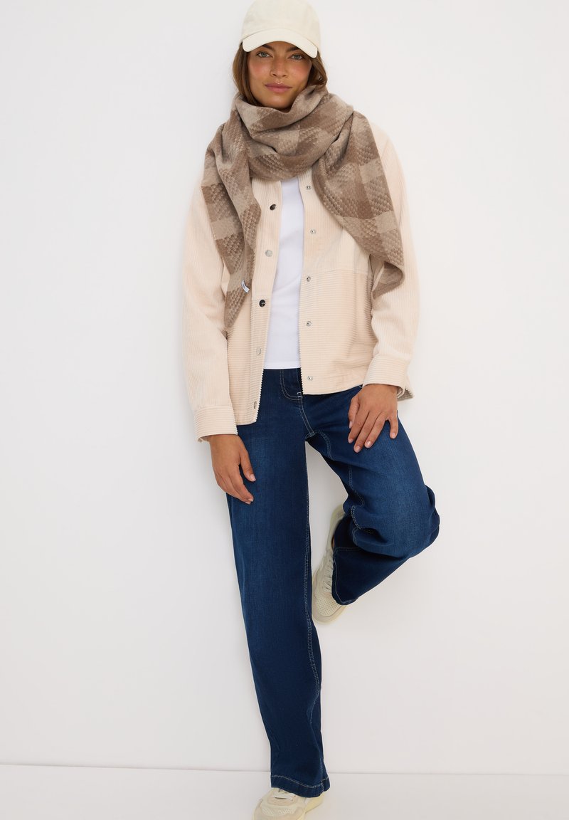 Donna con cappello bianco, giacca beige con bottoni, camicia bianca, grande sciarpa a quadri marrone, jeans blu e sneakers bianche, in piedi con una gamba sollevata.