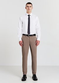 Jeune homme debout portant une chemise blanche, une cravate noire, un pantalon beige, une ceinture noire et des chaussures de ville noires devant un fond uni.