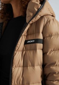Pufferjacke in tan met een geborduurd ontwerp, voorzien van een zwarte ritssluiting en een patch met "JACKIT" aan de linkerkant. Soepele textuur.