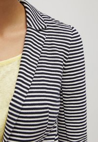 Gestreepte blazer in marineblauw en wit, met een gestructureerde stof, een getailleerde reverskraag en een borstzakje. Draagt over een geel topje.