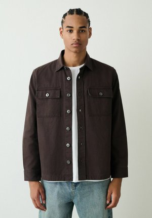 Dunkelbraunes Hemd aus Baumwolle mit Knöpfen, zwei Brusttaschen, silbernen Knöpfen und lässiger Passform über einem weißen T-Shirt.