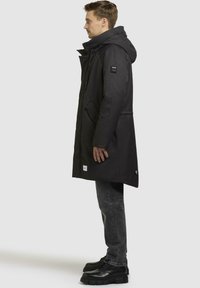 Sort parka med hætte, lynlås foran, sidelommer og et logo-patch på ærmet. Struktureret stof og en lang, løs pasform.