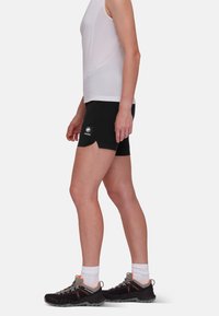 Black shorts with a side slit and logo patch, paired with a fitted white sleeveless top and black outdoor shoes with orange accents.