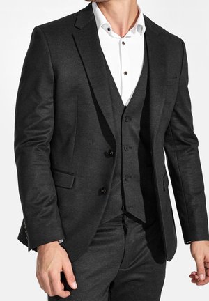Completo grigio scuro con revers a punta, giacca monopetto e pantaloni coordinati. Camicia bianca sotto con bottoni marroni.