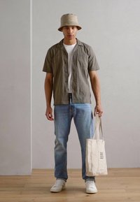Conjunto casual que incluye una camisa de manga corta verde oliva, una camiseta blanca, unos jeans azul claro, unas zapatillas blancas y una bolsa de playa beige.