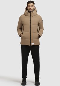 Beige pufferjack met een hoge kraag en kap, voorzien van een rits aan de voorkant, twee zijzakken en zwarte accenten bij de rits en de zoom.