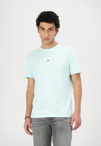 Hellblaue Baumwoll-T-Shirt mit Rundhalsausschnitt, kurzen Ärmeln und dem Logo-Text "TOMMY JEANS" in kontrastierenden Farben auf der Vorderseite. Getragen zu grauen Jeans.