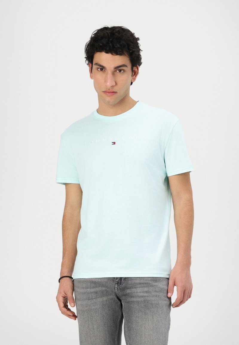 T-shirt in cotone azzurro chiaro con colletto tondo, maniche corte e logo frontale "TOMMY JEANS" con colori a contrasto. Indossato con jeans grigi.