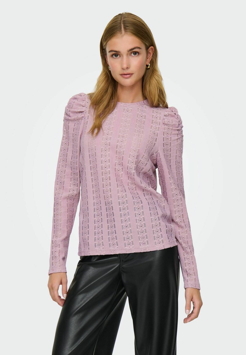 Kvinne med langt blondt hår som har på seg en lilafarget blonder topp med puffermer og sorte skinnbukser, stående foran en ensfarget bakgrunn.