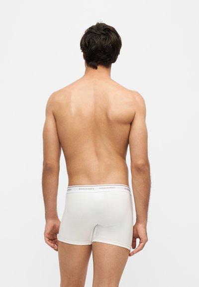 Homme debout de dos portant un boxer blanc avec le logo DSQUARED2 sur la ceinture, sur un fond gris uni.