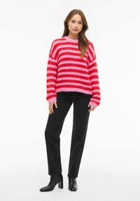 Różowo-czerwony sweter w paski, z miękkiego, puszystego materiału, z długimi rękawami, noszony na czarnych prostych spodniach i botkach na obcasie.