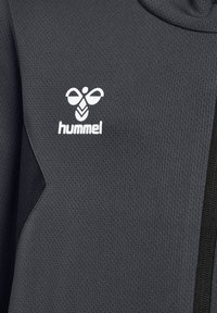 Nahaufnahme eines dunkelgrauen, strukturierten Stoffes mit weißem Hummel-Bienenlogo und Markennamen auf einer Sportjacke.