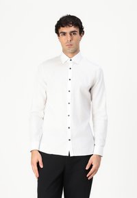 Hvit skjorte med lange ermer og spiss krage, med svarte knapper foran og rett kant. Glatt tekstur, tettsittende design.