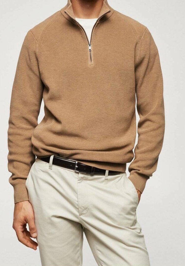 Homme portant un pull à demi-fermeture éclair beige par-dessus une chemise blanche, un pantalon beige et une ceinture noire, debout avec une main dans la poche.