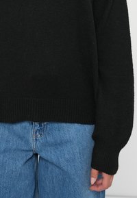 Svart strikket genser med ribbestrikket kant og mansjettdetaljer, sammen med lyseblå høyt liv denimjeans. Myk tekstur og avslappet passform.