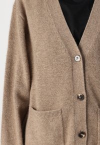 Beige svetr vyrobený z měkkého materiálu, s výstřihem do V, čtyřmi knoflíky a dvěma předními kapsami. Texturovaný povrch se jemným vzorem.