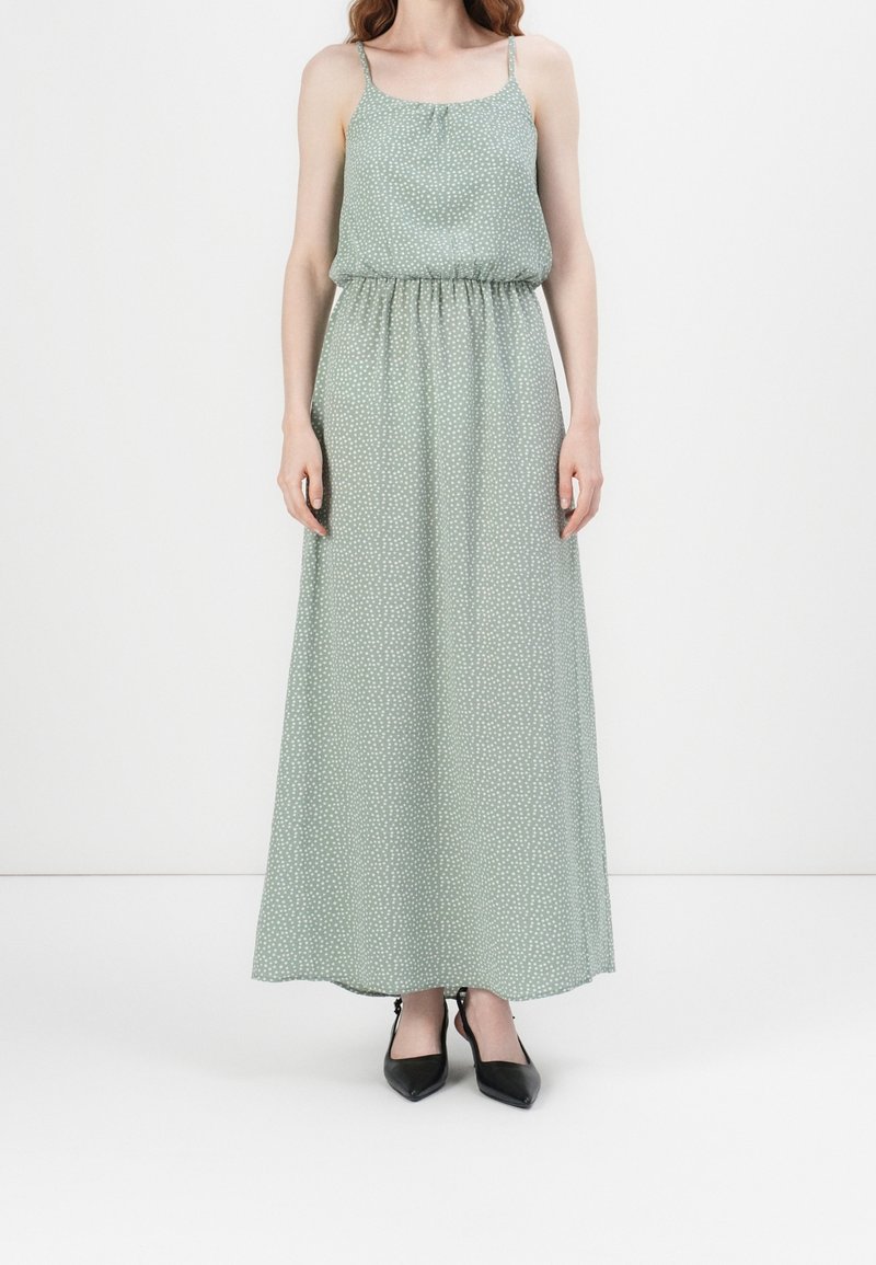 Femme portant une robe longue verte à pois blancs avec des fines bretelles, associée à des escarpins noirs à bout pointu.
