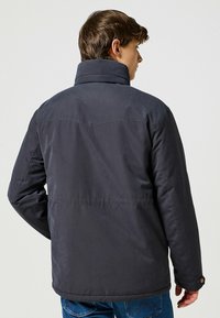 Chaqueta acolchada gris oscuro con un cuello alto, que presenta una textura suave, mangas largas y detalles de botones. Costuras visibles y un ajuste ajustado.