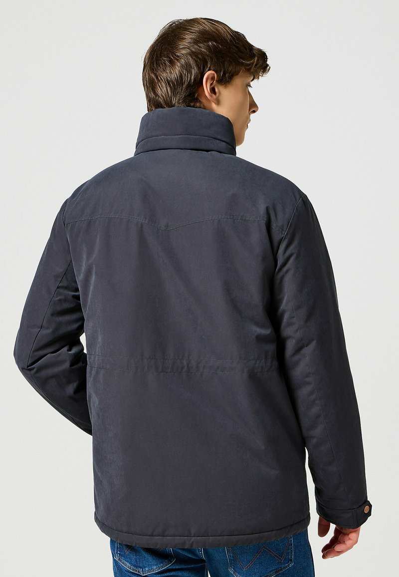 Giacca imbottita grigia scura con colletto alto, caratterizzata da una texture liscia, maniche lunghe e dettagli con bottoni. Cuciture visibili e vestibilità sartoriale.