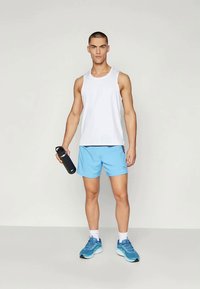 White athletic tank top, light blue shorts, and blue running shoes. Holding a black water bottle. Standing on a plain background.