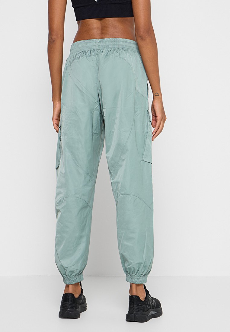 Person, die hellgrüne Cargo-Hosen mit elastischen Bündchen, schwarze Sneakers und ein schwarzes Cropped-Top trägt, steht vor einem weißen Hintergrund.