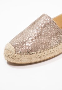 Gros plan d'une espadrille dorée rose pailletée avec des détails en jute tissée autour de la semelle et une surface supérieure texturée.