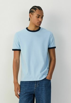 Mladý muž s opletenými vlasmi, oblečený v svetlomodrom tričku s tmavomodrým golierom a lemami rukávov a modrých džínsoch, stojaci s jednou rukou v vrecku.