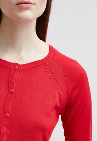 Close-up of a person wearing a red buttoned cardigan with ribbed stitching detail near the shoulder against a plain light background.