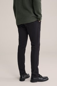 Černé kalhoty s úzkým střihem a hladkou texturou; doplněné lesklými černými mokasíny. Pozadí je neutrální béžová.