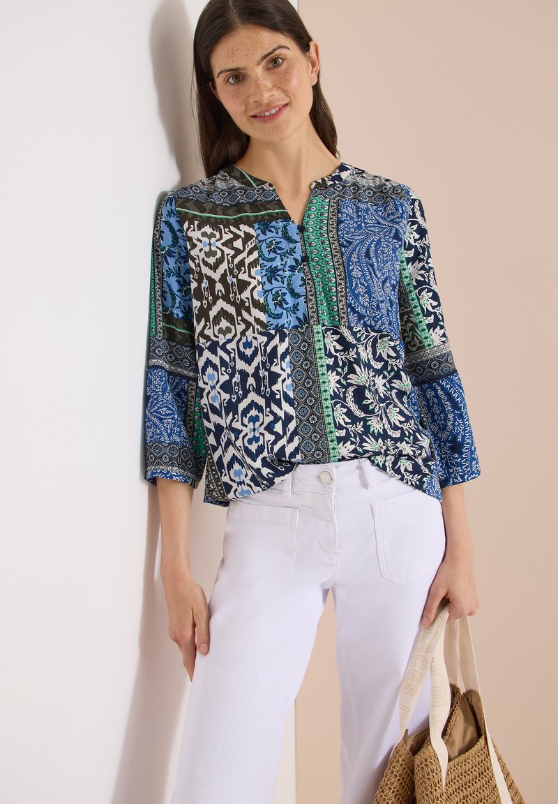 Femme portant un chemisier à motifs bleu, vert et noir avec un pantalon blanc, tenant un sac cabas tissé avec des poignées beige.