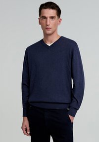 Bărbat îmbrăcat cu un pulover bleumarin în formă de V și pantaloni negri, stând cu o mână în buzunar pe un fundal gri uniform.