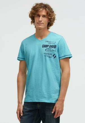 Young man with curly hair wearing a light blue V-neck T-shirt with dark text and graphics on the chest, paired with dark jeans.