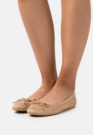 Femme portant des ballerines en suède beige avec des nœuds décoratifs, se tenant sur un fond blanc.