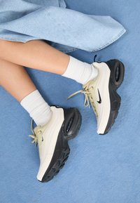 Nike Air Max tenisice imaju bež i crnu boju, s glatkim gornjim dijelom, vidljivom zračnom amortizacijom i ravnim gumiranim potplatima.