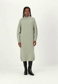 Φως πράσινο φόρεμα με μακριά μανίκια φτιαγμένο από μαλακό πλεκτό ύφασμα, με ψηλό λαιμόκοψη και ίσια σιλουέτα, συνοδευόμενο από μαύρες μπότες έως το γόνατο.