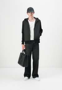 Crna zip-up majica s kapuljačom, bijela majica, crne široke traperice, bejzbol kapa i bijele patike; drži crnu torbu. Minimalistički dizajn.