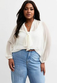 Weißes, durchsichtiges Blusenoberteil mit langen, luftig fallenden Ärmeln und einer gebundenen Vorderseite. Kombiniert mit hoch geschnittenen blauen Jeans. Sanfte Textur, leichtes Material.
