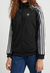 Svart zip-up jacka med vita randiga ärmar, med en liten Adidas-logotyp på bröstet och ribbade ärmslut för en säker passform.
