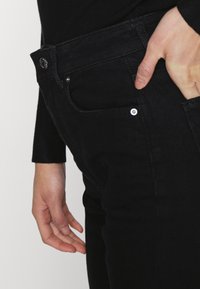 Jeans de ganga pretos com cintura média, bolsos frontais e um pequeno detalhe em botão de metal. O tecido tem uma textura suave.